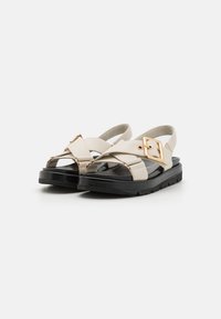 Sandales beige avec un design à lanières croisées, accent en boucle dorée, semelle en caoutchouc noir et semelle intérieure texturée. Talon plat pour un confort optimal.