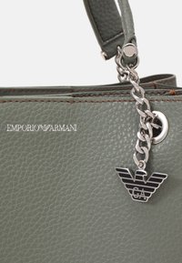 Zelená kožená kabelka s texturou, vybavená stříbrným řetízkem a přívěskem, s logem vystouplým na přední straně. Viditelný detail se zipovým uzávěrem.