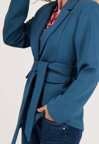 Teal blazer met een strikceintuur, brede mouwen en een gestructureerd ontwerp. Gecombineerd met een gebloemd blouse en donkere spijkerbroek.