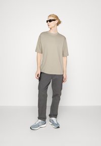 Lichtgrijs T-shirt, losse pasvorm, gecombineerd met donkergrijze cargo-broek met zijzakken. Schoeisel: blauw en witte sportschoenen, donkere zonnebril.