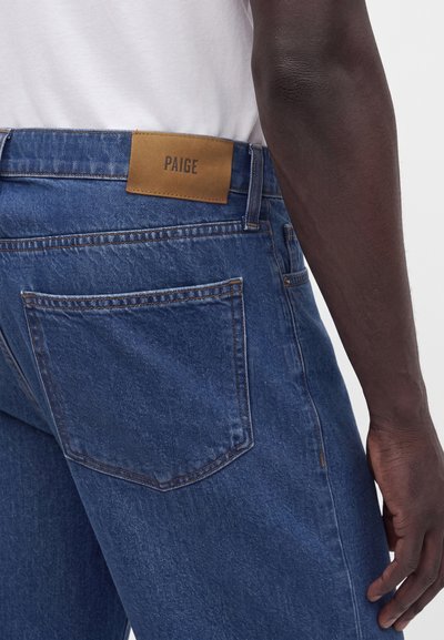 Κοντινό πλάνο ενός ατόμου που φορά μπλε τζίν denim με καφέ δερμάτινη ετικέτα με την ένδειξη "PAIGE" στη μέση και ένα λευκό πουκάμισο μέσα.