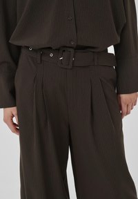 Tummanruskeat suorat housut ja niihin matching-paita, leveä vyö ja neliönmuotoinen solki, henkilö, jonka kädet ovat rentona sivuilla.