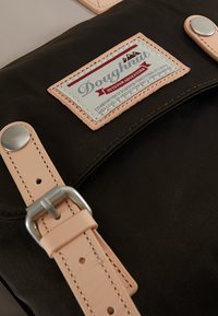 Gros plan d’un sac à dos noir avec des sangles en cuir beige et une étiquette Doughnut Outdoor Adventure cousue sur le rabat avant.