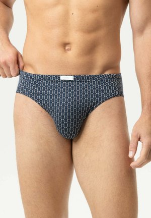 Torse masculin portant un slip bleu marine à motifs, ajustant la ceinture d'une main.
