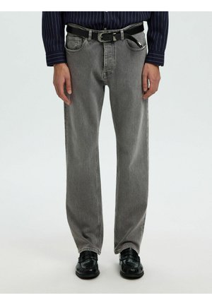 Homme portant un jean gris ample avec une ceinture noire, des chaussures noires texturées et une chemise rayée bleu foncé avec des manches visibles jusqu'aux poignets.