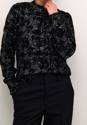 Personne portant un haut à manches longues noir à sequins associé à un pantalon noir, une main dans la poche, l'autre touchant le cou.