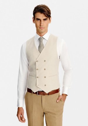 Mężczyzna noszący beżową dwurzędową kamizelkę, białą koszulę, wzorzysty krawat, brązowy pasek i jasnobrązowe spodnie, stojący z jedną ręką w kieszeni.