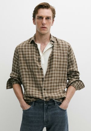 Giovane uomo con le mani in tasca che indossa una camicia a quadri marrone sopra una henley bianca e jeans blu scuro su uno sfondo chiaro e semplice.
