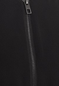 Zwarte zip-up jas van een gladde, gestructureerde stof. Beschikt over een metalen rits met een rechthoekige trekker, die verticaal aan de voorkant loopt.