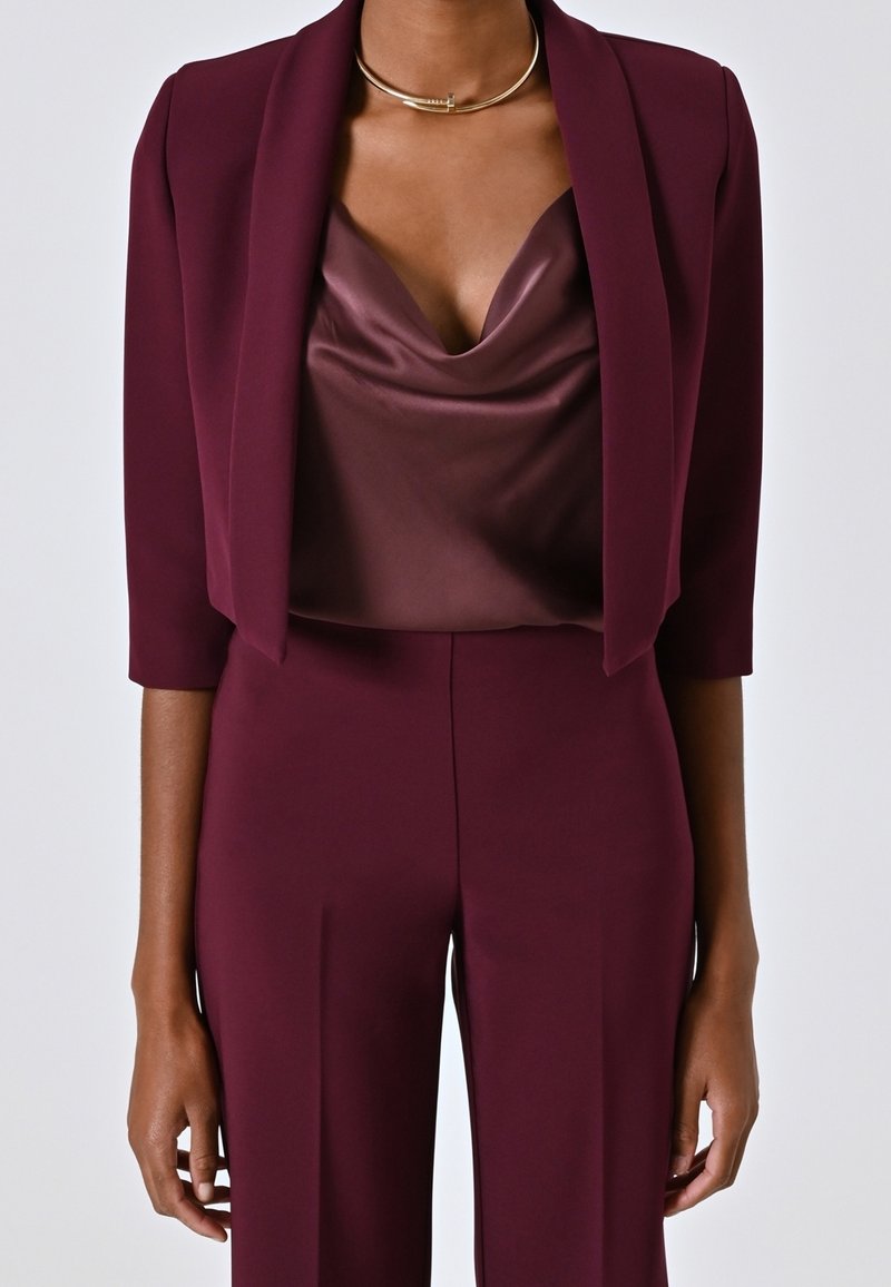 Femme portant un blazer court bordeaux, un top drapé en satin, un pantalon taille haute assorti et un collier ras-du-cou doré.