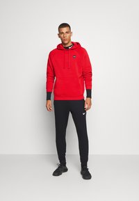 Rode hoodie met een voorzak, gecombineerd met zwarte joggingsbroek met een wit Nike-logo. Beide bieden een casual sportieve uitstraling.