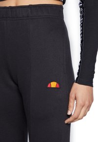 Svarta mjukisbyxor med resår i midjan, som har en liten broderad logotyp i rött, orange och gult på vänstra låret.