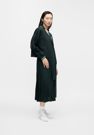 Femme portant un haut à manches longues vert foncé avec des bordures blanches, une jupe midi plissée, des chaussettes blanches et des baskets blanches, debout devant un fond uni.