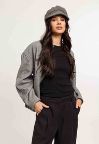Grau melierte Wolljacke mit übergroßen Ärmeln, kombiniert mit einem schwarzen, taillierten Oberteil und schwarzen, gefältelten Hosen. Eine Kappe mit Fischgrätenmuster rundet den Look ab.