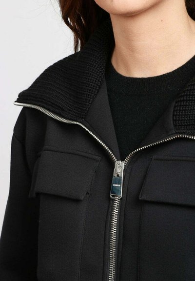 Giacca zip-up nera realizzata in tessuto morbido, con un colletto a maglia testurizzata, due tasche frontali e una cerniera metallica con un tirante circolare.