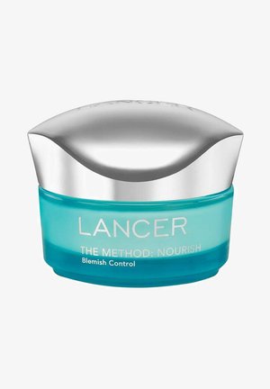 Een ronde, turquoise glazen pot met een zilveren, gebogen deksel. Bevat de tekst "DE METHODE: VOEDEN Blemish Control" in het wit op de voorkant.