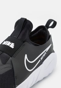 Czarne buty sportowe typu slip-on z teksturowanym wierzchem, elastycznym paskiem z logo "Nike", białym akcentującym logo oraz białą gumową podeszwą.