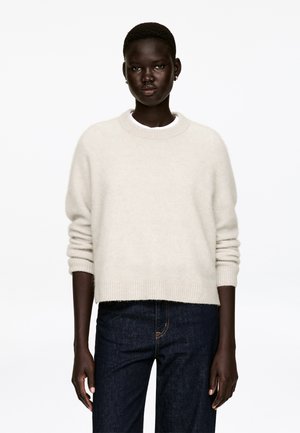 Hellbeiger Pullover aus weichem Strickmaterial, mit Rundhalsausschnitt, langen Ärmeln und lockerem Schnitt, kombiniert mit dunklen Jeans aus Denim.