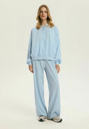 Młoda kobieta stoi, ubrana w jasnoniebieską kurtkę na zamek i szerokie spodnie, ma na sobie szare trampki, na jednolitym tle.