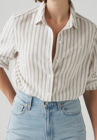 Chemise blanche à rayures avec fermeture à boutons, présentant des lignes verticales en violet et bleu. Tissu doux et léger, coupe oversize et manches courtes.