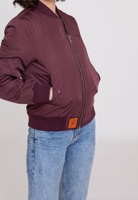 Giacca bomber bordeaux con zip frontale, polsi a coste e patch con logo sull'orlo. Abbinata a jeans in denim azzurri.