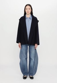 Cappotto in lana oversize blu navy con ampi rever, abbinato a una camicia azzurro chiaro e a un paio di jeans blu sbiaditi e larghi. Ballerine nere completano l'outfit.