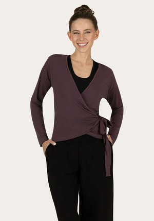 Femme souriante avec les cheveux en chignon porte un haut cache-cœur à manches longues violet foncé noué à la taille sur une chemise noire et un pantalon noir, les mains dans les poches.