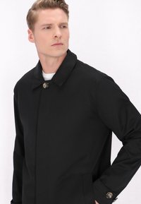 Impermeabile nero con colletto, chiusura a un bottone e due tasche laterali. Tessuto liscio e resistente all'acqua. Accenti di hardware lucido.