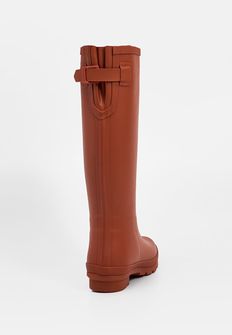 Bota de lluvia de goma marrón con un diseño alto y plano. Presenta una suela con agarre texturizado y un detalle de hebilla en la parte superior trasera para ajuste.