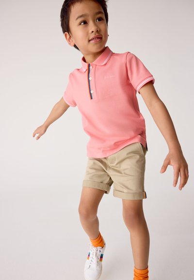 Roze poloshirt met contrasterende kraag en knopen, gecombineerd met beige shorts, oranje sokken en witte sneakers met kleurrijke accenten.