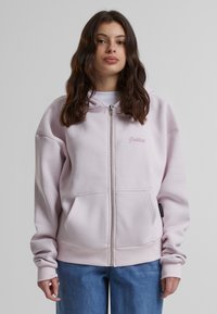 Felpa con zip rosa chiaro, realizzata in tessuto morbido, con tasca frontale, polsini a costine e un logo ricamato sul petto in modo discreto.