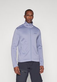 Šviesiai purpurinė zip-up striukė su aukštu apykakle, tekstūruotas audinys ir šoniniai kišeniai, derinama su tamsiomis krovinių šortais; logotipas viršuje kairėje pusėje.