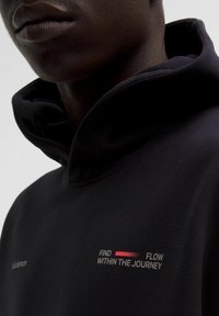 Nahaufnahme einer Person, die einen schwarzen Kapuzenpulli trägt mit dem Text "Finde Fluss innerhalb der Reise" und einem kleinen Lululemon-Logo auf der Brust.