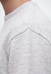 Gros plan sur l'épaule et le haut de la manche d'un pull en maille gris clair avec des détails de coutures visibles sur une personne de profil.