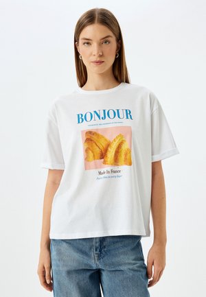 Camiseta blanca de manga corta con un estampado gráfico de croissants. El texto en azul y rosa dice "BONJOUR" y "Hecho en Francia". Corte casual.