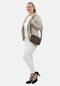 Læderet crossbody taske med leopardmønster, sort hardware, rektangulær form og tekstureret stof. Modellen bærer en lys beige jakke og hvide bukser.
