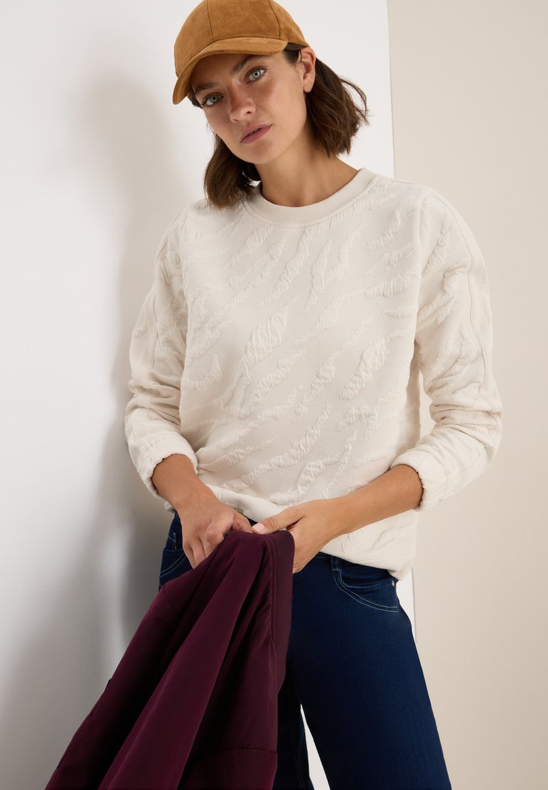 Pull en beige clair texturé avec col rond, associé à un jean bleu foncé. La personne tient une veste bordeaux. Une casquette marron complète la tenue.