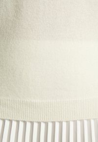 Ourlet d'un pull en maille couleur crème au-dessus d'une jupe plissée blanche, montrant la texture et les détails du tissu dans des tons doux et neutres.