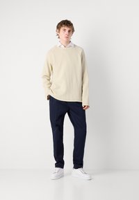 Beige langermet genser med rund hals, marineblå smale bukser og hvite joggesko. Genseren har et teksturert stoff; buksene har sidelommer.