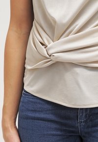 Beige, slät tygöverdel med en tvinnad knutdetalj vid midjan, kombinerad med mörkblå denimjeans. Texturen är mjuk och elastisk.