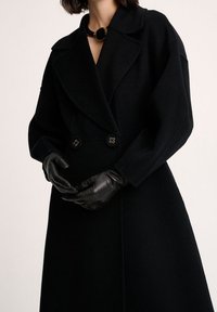 Czarny wełniany płaszcz z dwurzędowym zapięciem, dużymi klapami i ozdobnymi guzikami, w połączeniu z czarnymi skórzanymi rękawiczkami.