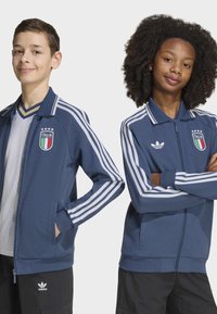 Dvaja tínedžeri oblečení v modrých futbalových bundách Adidas Italy s pásikmi stoja sebavedomo pred bielym pozadím.