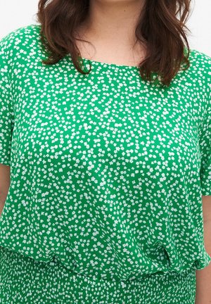 Femme portant une robe verte à petits motifs floraux blancs, manches courtes et taille froncée, cheveux châtains mi-longs.
