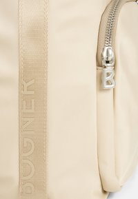 Béžový nylonový batoh s textúrovaným popruhom a logom "BUGNER", striebornými kovovými detailmi a zipsovou kapsou. Hladký, čistý povrch.