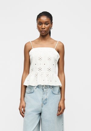 Ung kvinde med flettet hår iført hvid ærmeløs øjenlet top og lyseblå højtaljede denimjeans, stående mod ensfarvet baggrund.