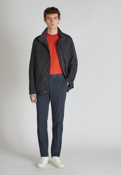 Giacca leggera nera con chiusura a bottoni a pressione, dotata di due tasche frontali. Indossata sopra una camicia rossa e pantaloni blu marino, con sneakers bianche.