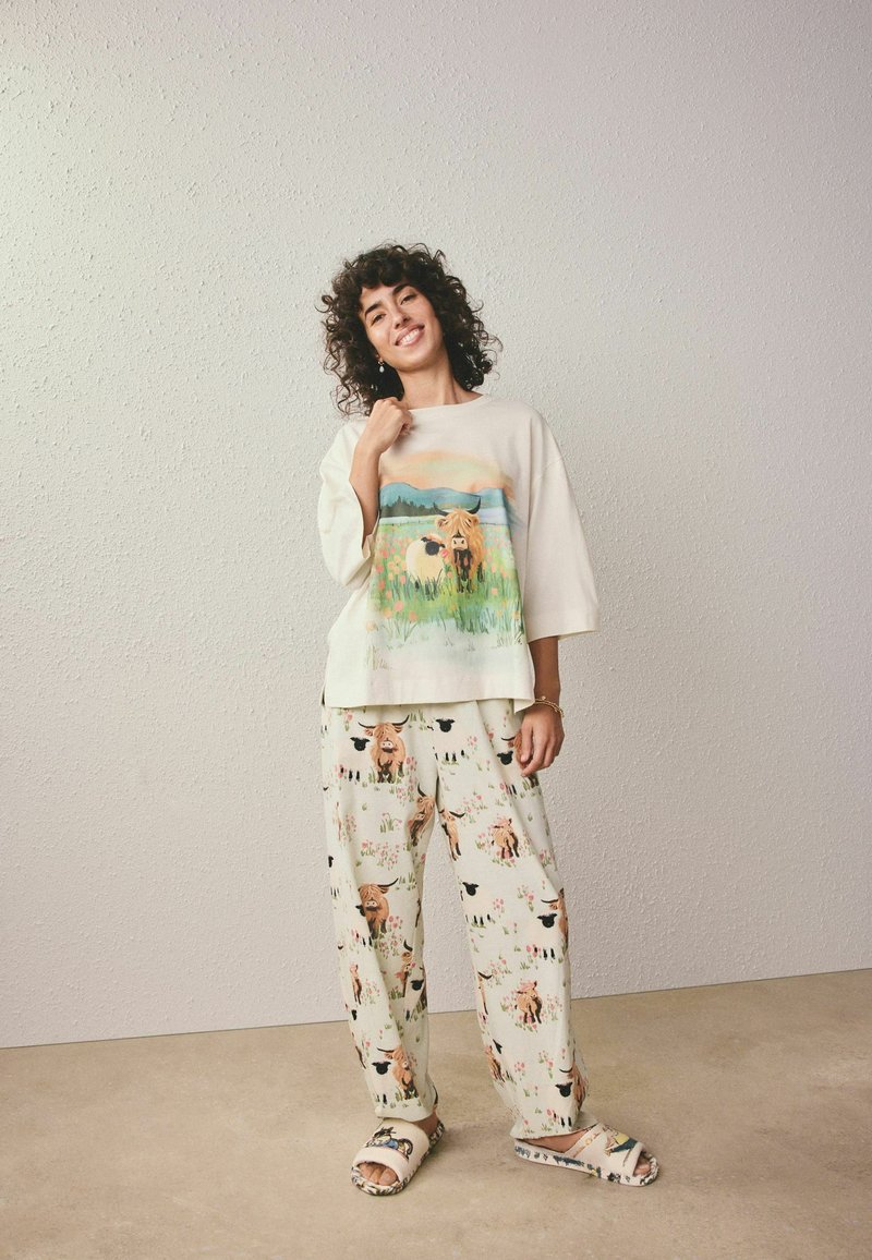 Femme en pyjama crème avec des illustrations de vaches, debout et souriante devant un mur texturé, portant des chaussons assortis.