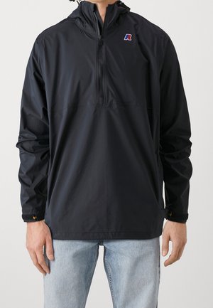 Muž v černé větrovce s kapucí a polovičním zipem s malým logem na hrudi a světlých modrých džínách stojí před jednobarevným pozadím.