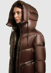 Barna puffdzseki magas kapucnival, fényes felülettel, steppelt textúrával és fekete cipzárokkal. Két cipzáras elülső zsebbel rendelkezik.