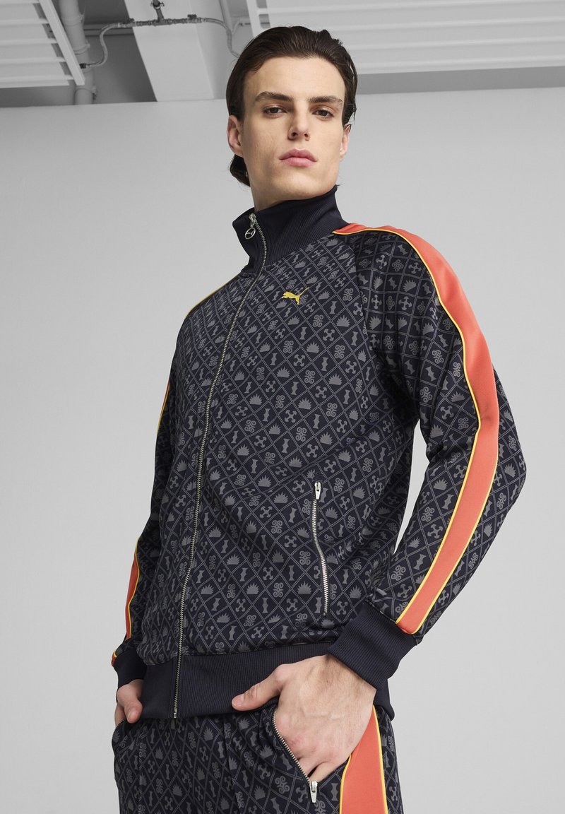 Giacca a track con motivo geometrico blu navy, strisce rosse e gialle, tasche con zip e colletto rialzato. Presenta un logo sul petto.